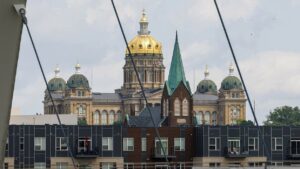 the-golden-dome-of-the-iowa-state-capitol-is-visible-through-wire-slats-on-a-suspension-bridge-over-a-river-in-front-of-the-cap